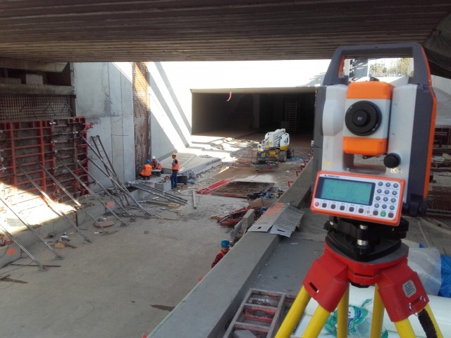 Tachimetr na statywie na tle placu budowy pod wiaduktem, widoczni pracownicy budowlani w pomarańczowych kamizelkach i kaskach.