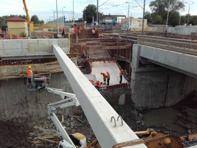 Budowa wiaduktu kolejowego: pracownicy w kaskach nadzorują wznoszenie betonowej konstrukcji, widoczny wysięgnik dźwigu i wykop z wodą.