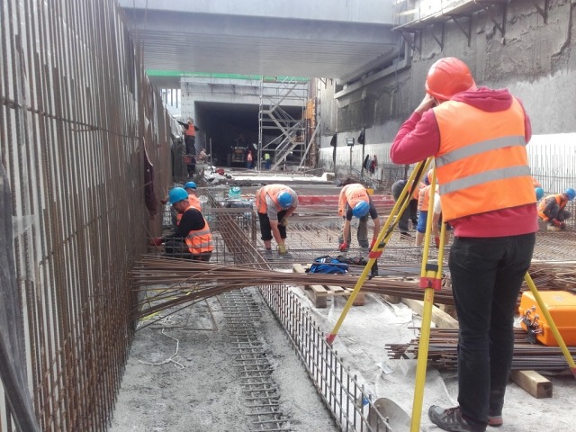 Prace budowlane przy budowie tunelu: pracownicy w kaskach i kamizelkach odblaskowych montują zbrojenie, a inżynier z pomarańczowym kasku obsługuje niwelator na statywie.