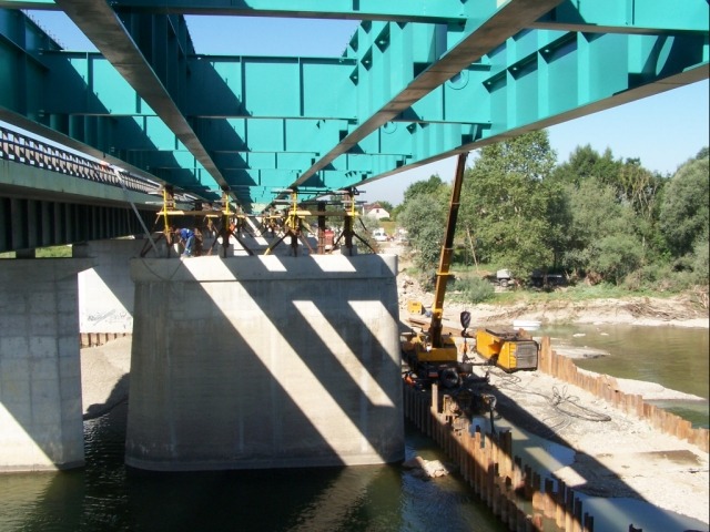 Budowa mostu nad rzeką: widoczne stalowe elementy konstrukcyjne pomalowane na turkusowo, betonowe filary, dźwig i robotnicy w oddali.