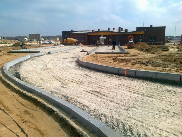 Budowa drogi dojazdowej z ułożonymi obrzeżami betonowymi, widoczne ślady opon na piasku i budynek w tle.