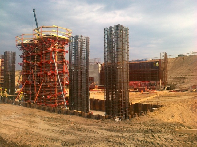 Budowa filarów mostu: szalunki i zbrojenie stalowe na placu budowy, widoczni pracownicy w żółtych kaskach.