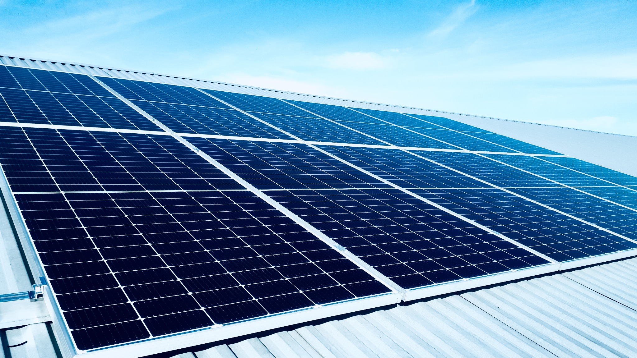 Panel słoneczny na falistym dachu budynku przemysłowego, błękitne niebo w tle.