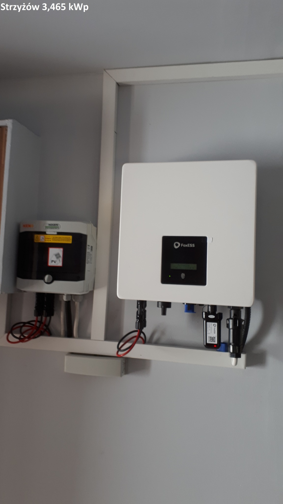Dwa inwertery fotowoltaiczne, jeden marki Nomoik z naklejką ostrzegawczą PV, drugi marki FoxESS, zamontowane na białej półce wewnątrz pomieszczenia, z widocznymi przewodami i złączami.