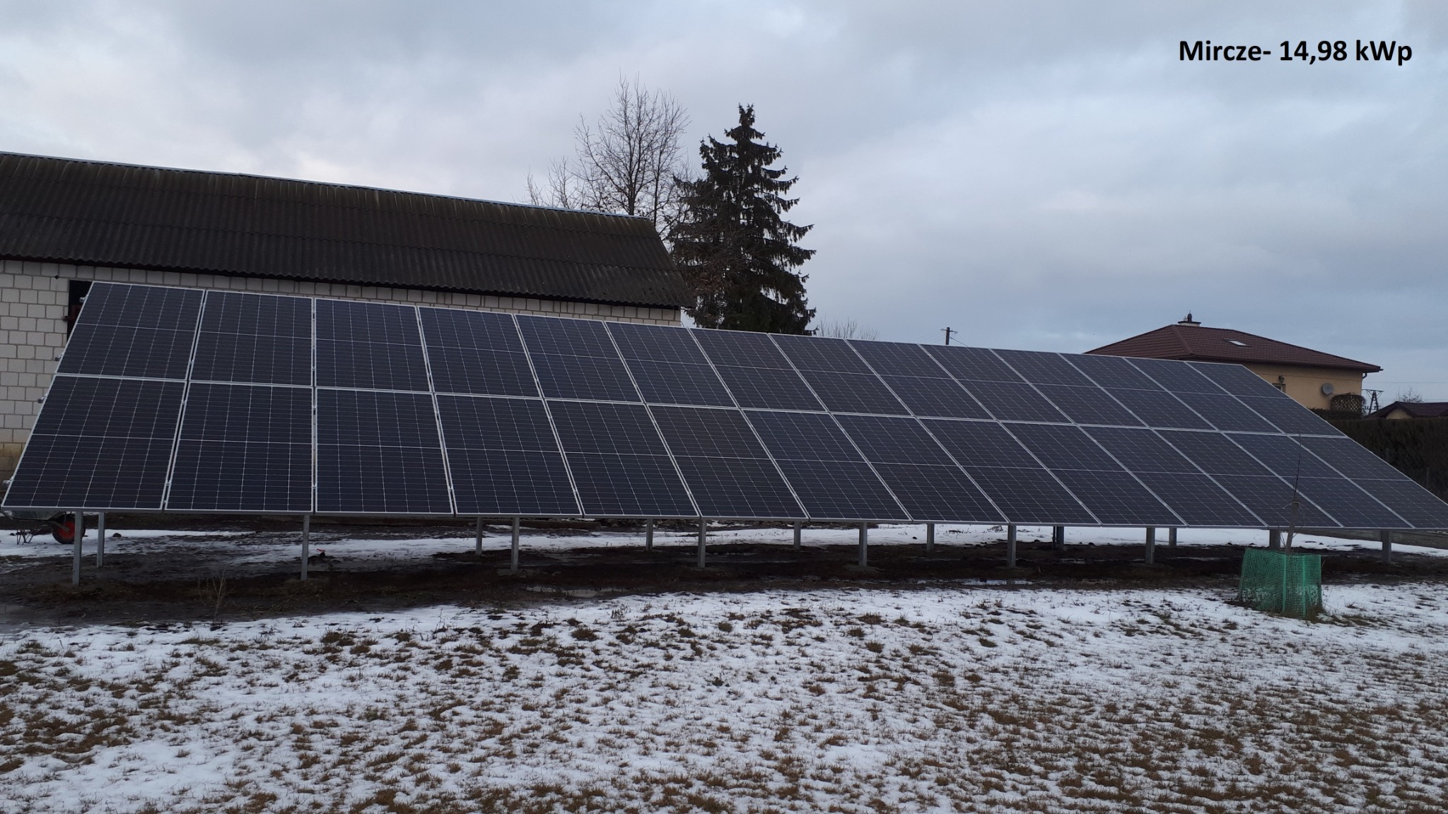 Instalacja paneli słonecznych o mocy 14,98 kWp na metalowej konstrukcji, z widocznym budynkiem gospodarczym w tle i resztkami śniegu na ziemi.
