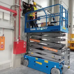 Dwóch monterów w kaskach i kamizelkach na podnośniku Genie GS-4047, instaluje okablowanie w szafie elektrycznej wewnątrz hali przemysłowej, widoczne rury instalacji przeciwpożarowej.