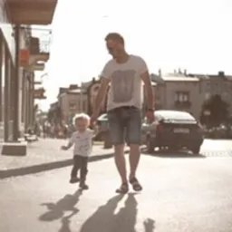 Ojciec trzymający za rękę małego syna na ulicy w słoneczny dzień, rzucają długie cienie.