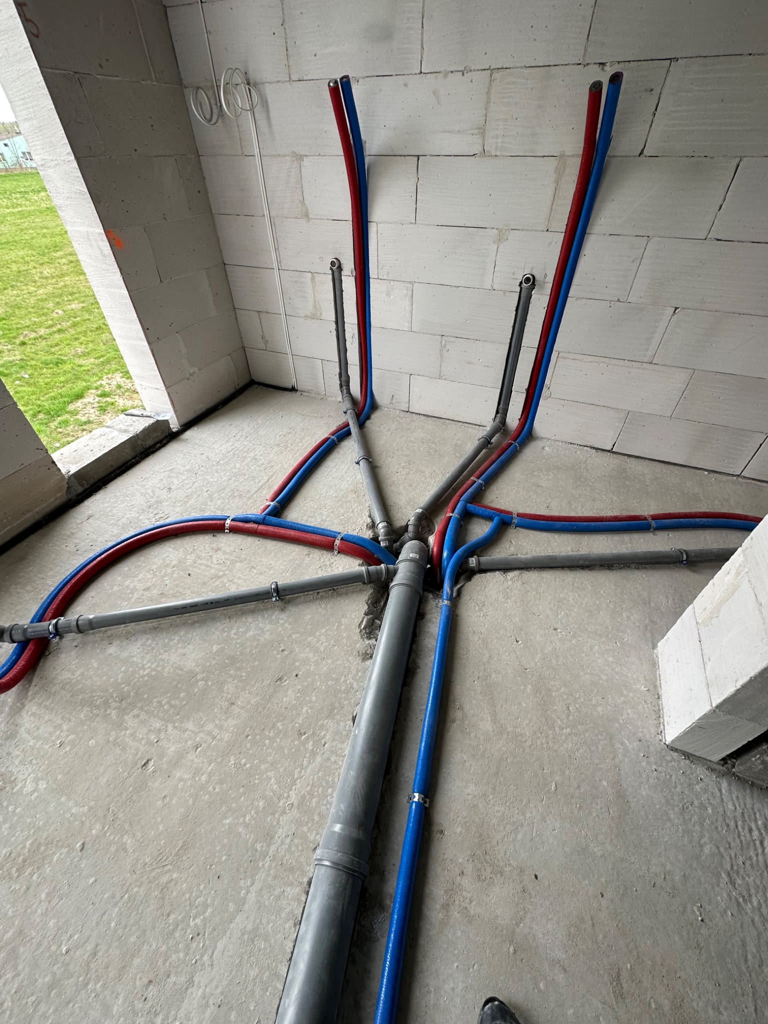 Rozprowadzenie rur wodnych i kanalizacyjnych w surowym stanie budynku, widoczne rury czerwone, niebieskie i szare, mocowane do podłoża za pomocą obejm.