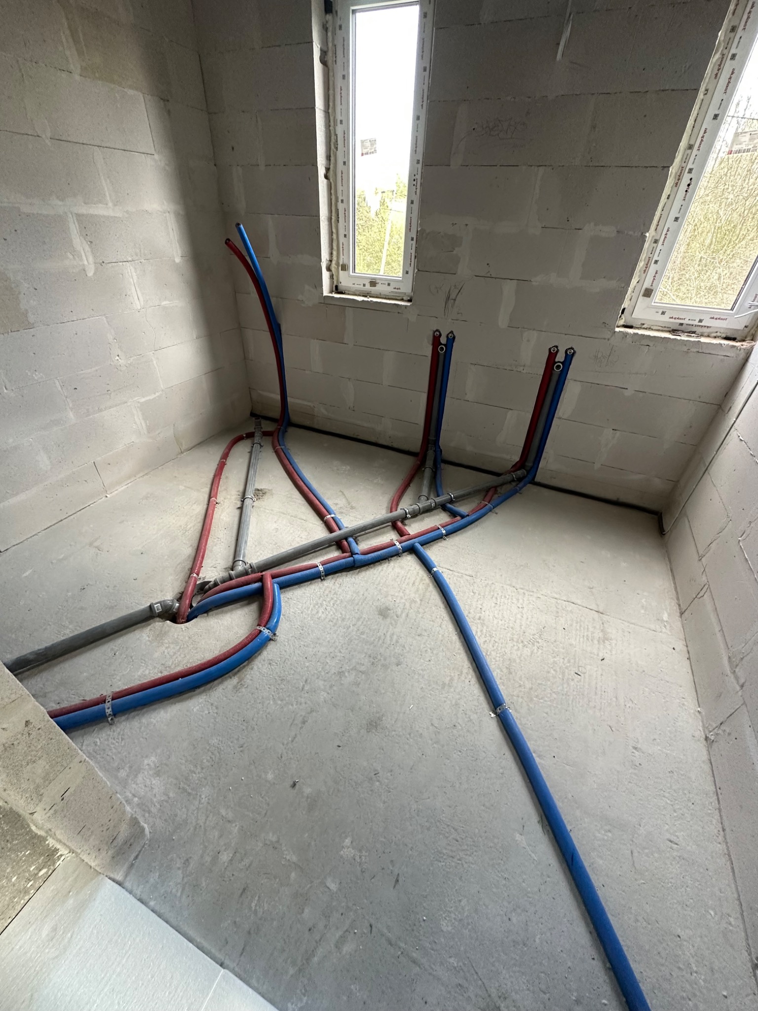 Surowy stan: Instalacja wodno-kanalizacyjna w pomieszczeniu z pustaków, widoczne rury PEX w kolorze czerwonym i niebieskim oraz szare rury kanalizacyjne prowadzone po podłodze i w górę w kierunku...