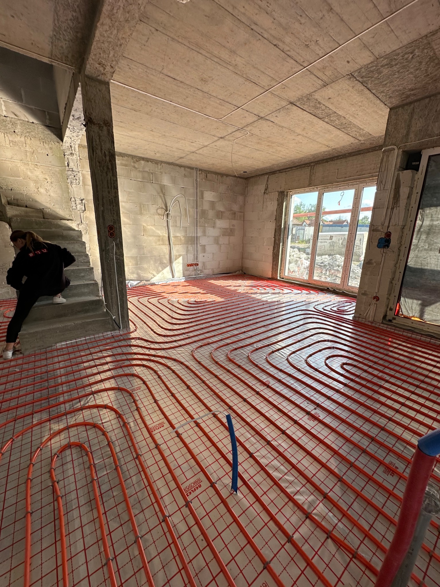Rozłożone czerwone rury ogrzewania podłogowego na folii w surowym wnętrzu domu w trakcie budowy, widok na okno i betonowe schody z osobą.