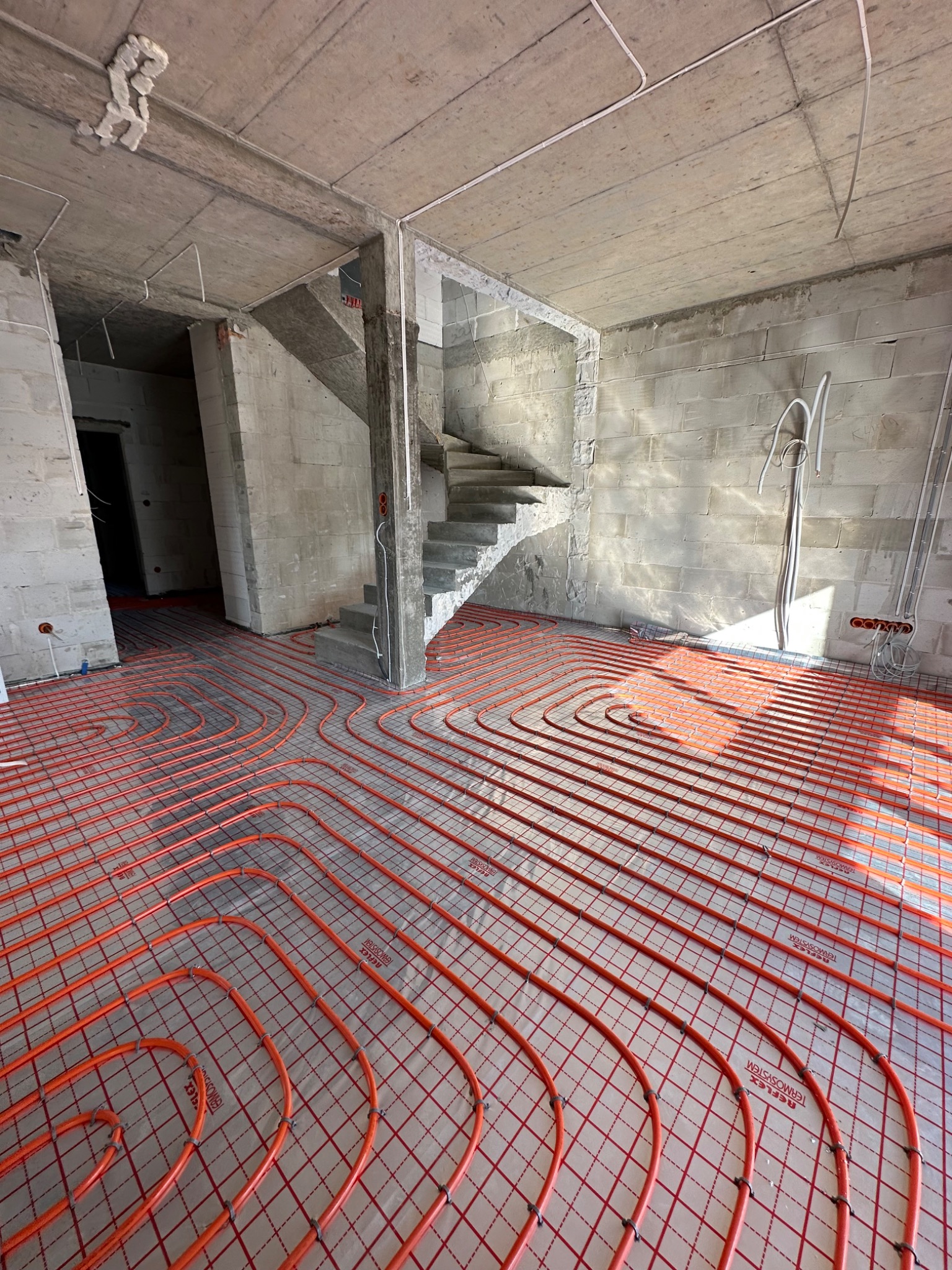 Pomarańczowe rury ogrzewania podłogowego ułożone na siatce w surowym wnętrzu z betonowymi ścianami i schodami, widoczne przewody elektryczne pod sufitem.