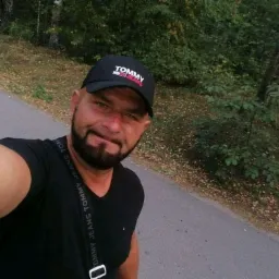 Selfie mężczyzny w czarnej koszulce i czapce Tommy Jeans na tle leśnej drogi.