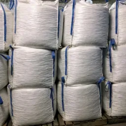 Stos białych work&oacute;w typu Big Bag z niebieskimi uchwytami, ułożonych na drewnianej palecie w magazynie.