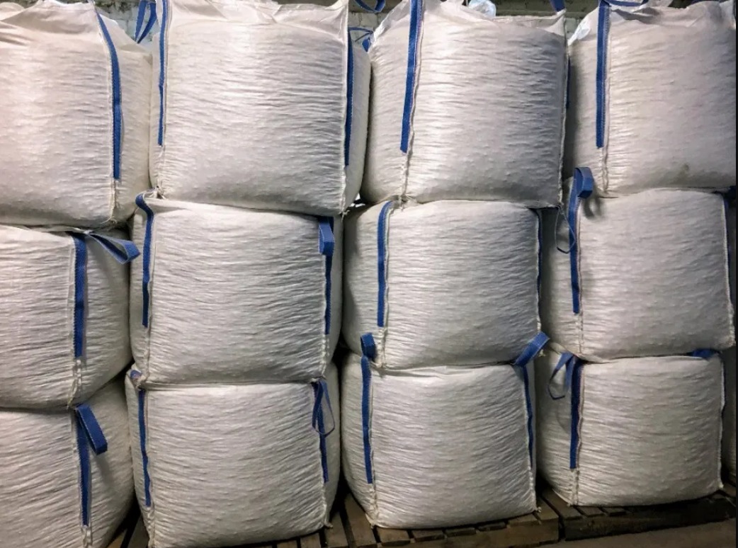 Stos białych worków typu Big Bag z niebieskimi uchwytami, ułożonych na drewnianej palecie w magazynie.