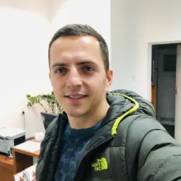 Uśmiechnięty mężczyzna w kurtce The North Face z żółtymi detalami na tle biura z drukarką, rośliną doniczkową i częściowo widocznym plakatem na ścianie.