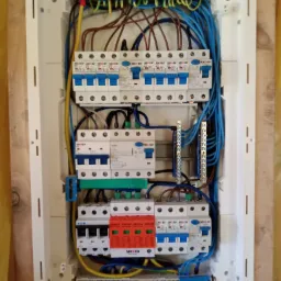 Otwarta skrzynka rozdzielcza z zamontowanymi bezpiecznikami, przewodami w kolorach niebieskim, brązowym i żółto-zielonym, widoczne listwy zaciskowe i ograniczniki przepięć.