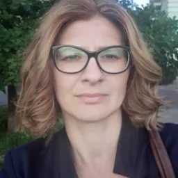 Portret kobiety w okularach i ciemnej marynarce na tle zieleni miejskiej, z delikatnym uśmiechem i profesjonalnym wyrazem twarzy.