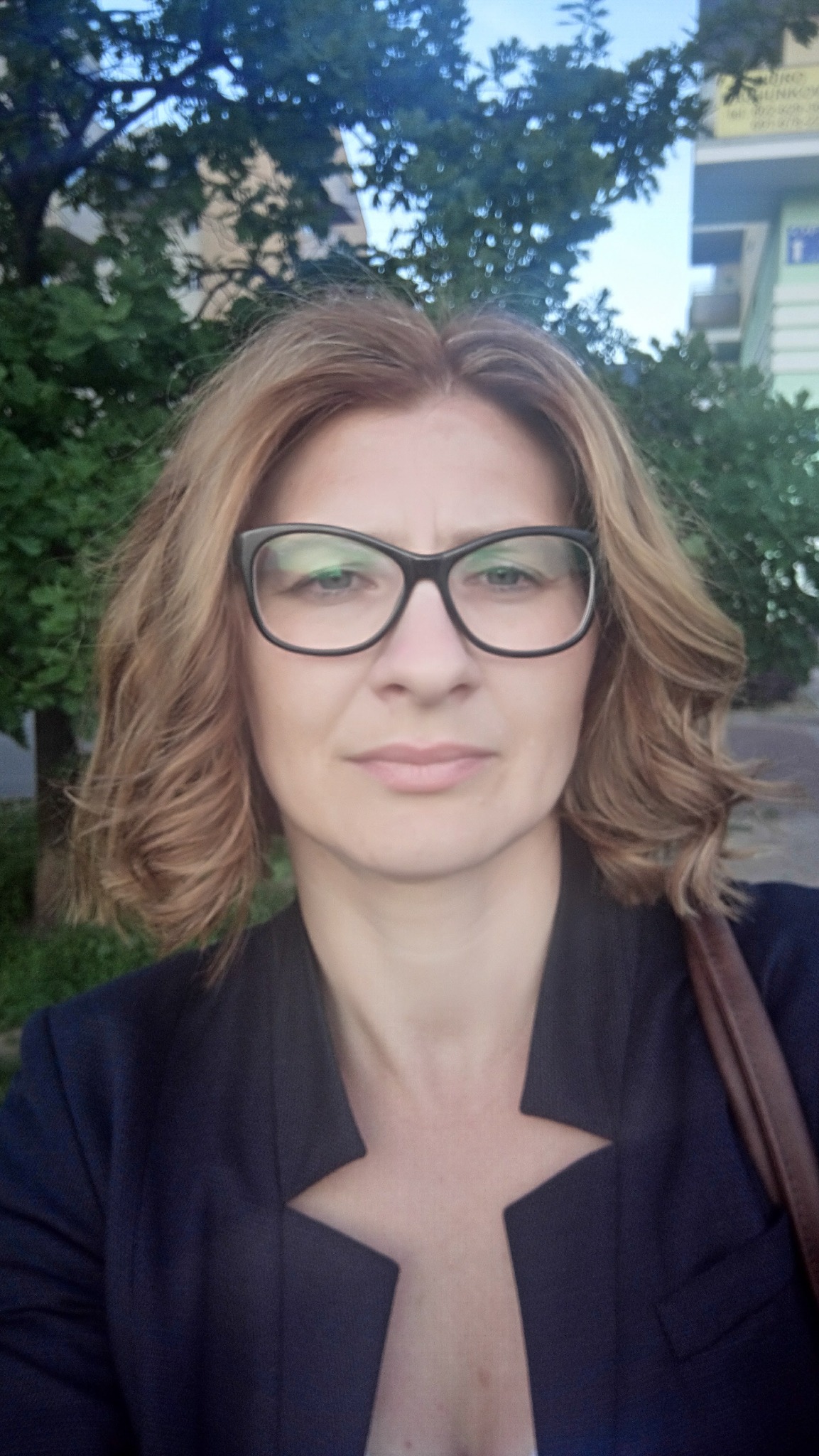 Portret kobiety w okularach i ciemnej marynarce na tle zieleni miejskiej, z delikatnym uśmiechem i profesjonalnym wyrazem twarzy.