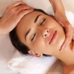 Kobieta z zamkniętymi oczami podczas masażu głowy i szyi, wykonywanego przez masażystę w jasnym otoczeniu.