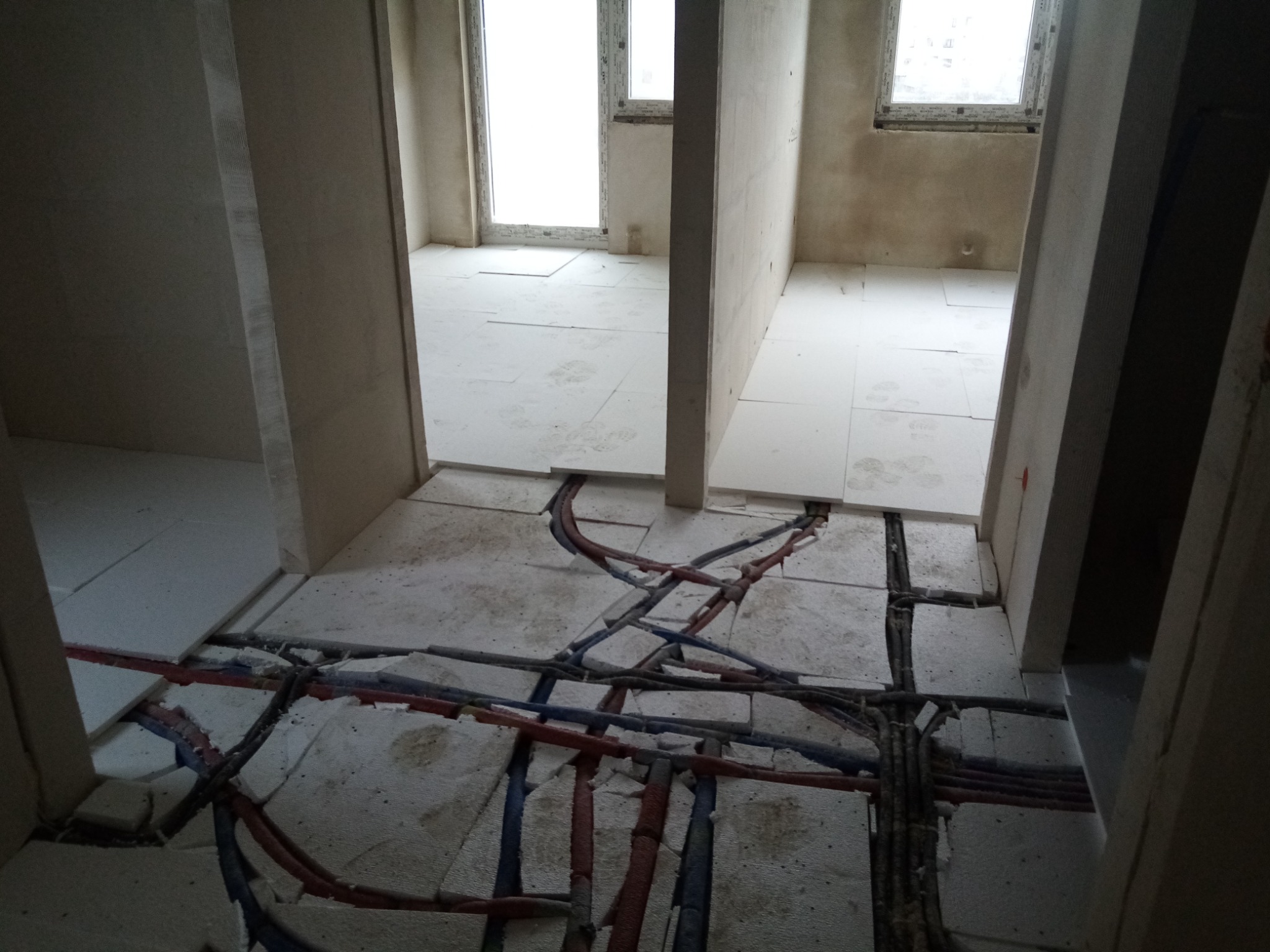 Przygotowanie podłoża pod posadzkę: izolacja styropianowa z rozłożonymi instalacjami wodno-kanalizacyjnymi w surowym stanie, widok z korytarza na dwa pomieszczenia.