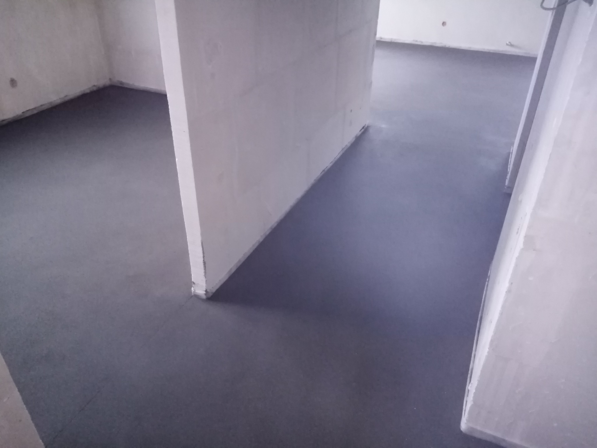 Szara, gładka posadzka w surowym wnętrzu z białymi ścianami w trakcie remontu, widoczne przejścia między pomieszczeniami.