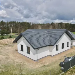 Nowoczesny, parterowy dom z keramzytu z grafitową dachówką, widok z góry na działkę w trakcie zagospodarowania, zalesione tło i pochmurne niebo.