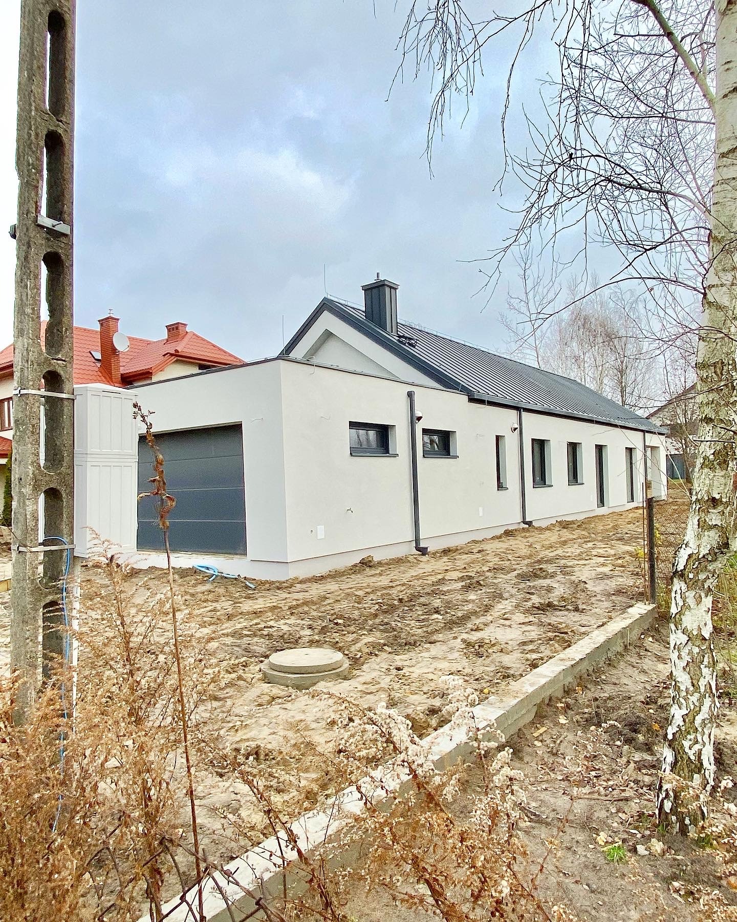 Niski, parterowy dom z garażem w stanie surowym, z szarą dachówką, widoczny słup elektryczny i sąsiedni dom z czerwoną dachówką w tle.