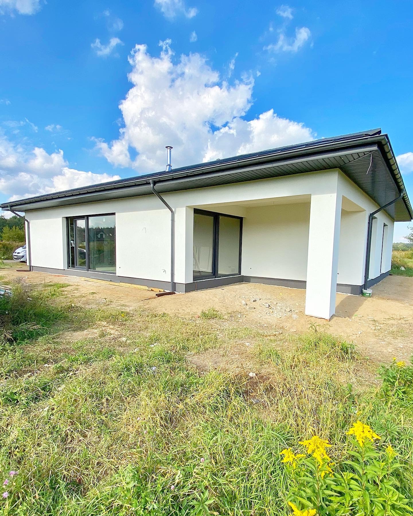 Parterowy, biały dom z ciemną stolarką okienną i zadaszonym tarasem, widoczny z zewnątrz na tle błękitnego nieba z chmurami i zarośniętej działki.