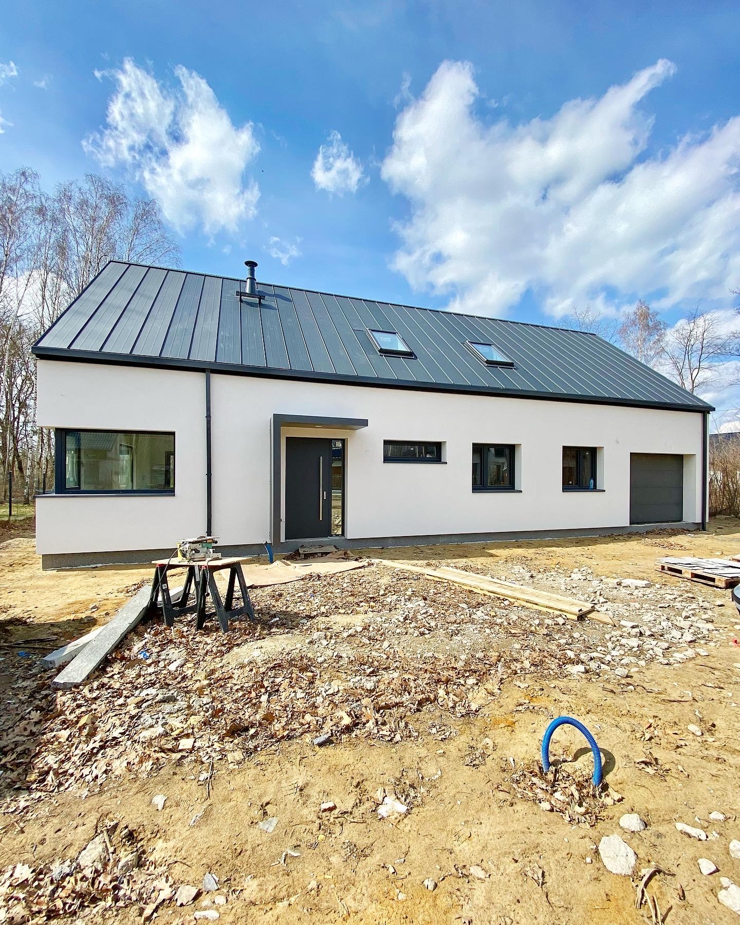 Nowoczesny dom jednorodzinny z szarą blachodachówką, widok z zewnątrz na plac budowy z piłą stołową i niebieskim peszelem wystającym z ziemi.