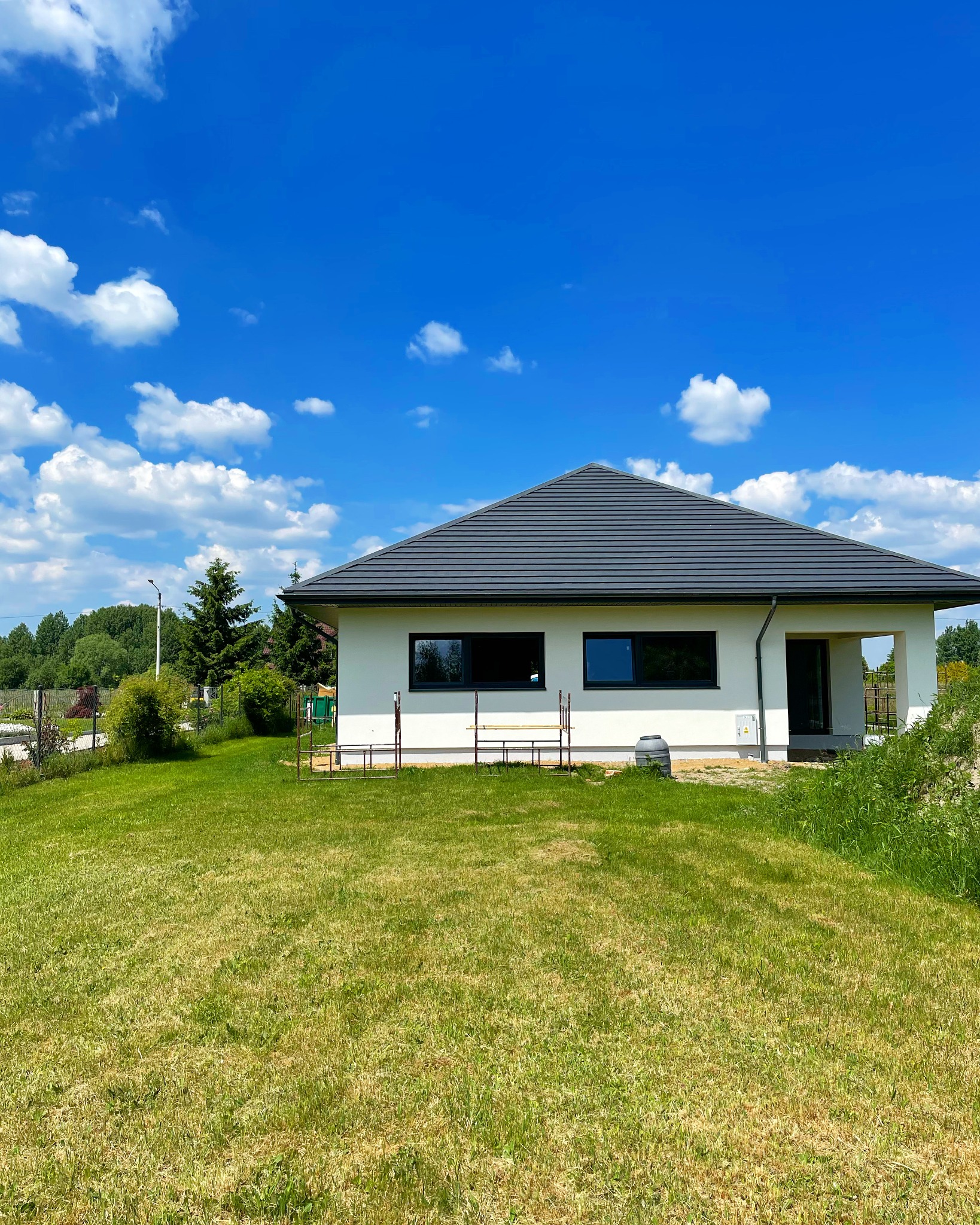 Biały parterowy dom z szarą dachówką, widok z trawnika w słoneczny dzień z błękitnym niebem i białymi chmurami, widoczne rusztowania i zbiornik na deszczówkę.