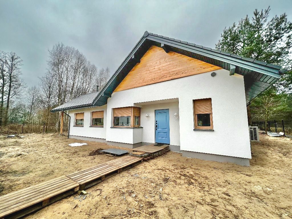 Niedawno wybudowany biały dom parterowy z niebieskimi drzwiami wejściowymi i drewnianymi okiennicami, otoczony piaszczystym terenem i drewnianym chodnikiem.