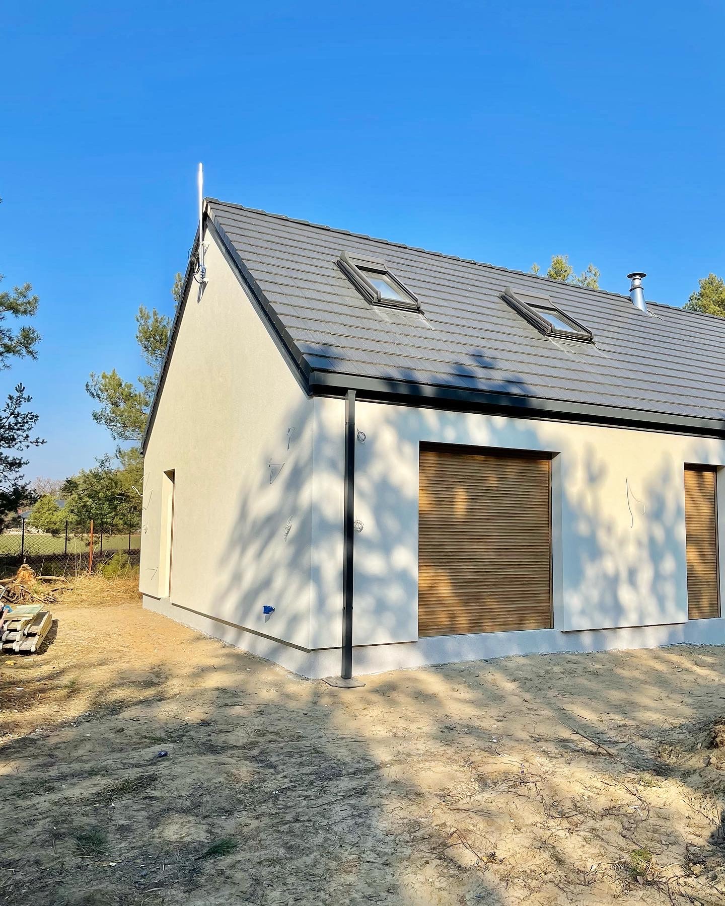 Nowoczesny dom jednorodzinny z szarą dachówką, dwoma oknami dachowymi i roletami zewnętrznymi, widziany z zewnątrz w słoneczny dzień.