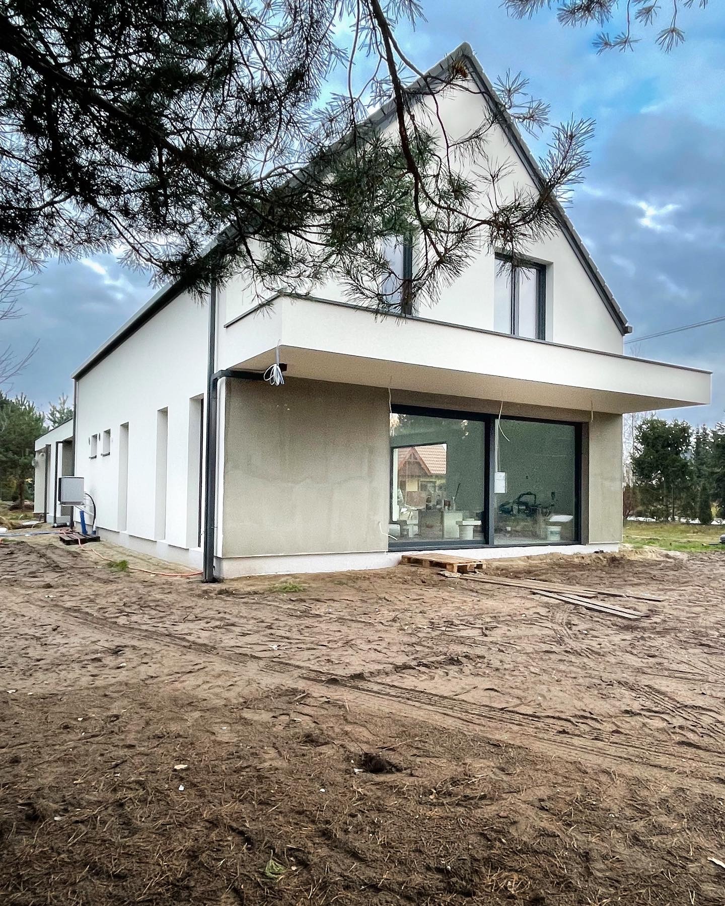 Nowoczesny dom jednorodzinny z dużym oknem tarasowym, widok z zewnątrz na plac budowy w trakcie prac wykończeniowych.