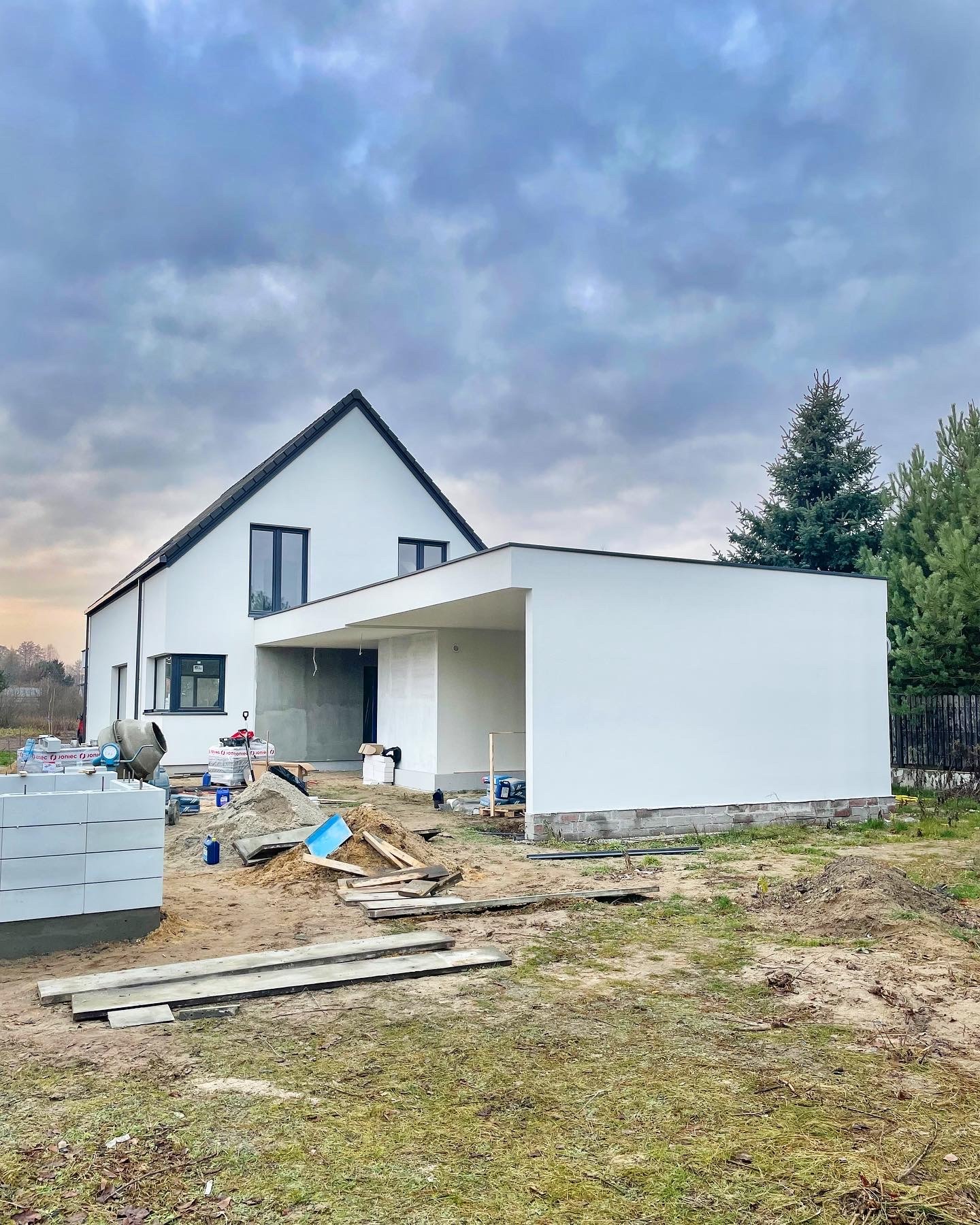 Nowoczesny, biały dom jednorodzinny w trakcie budowy, widoczny betonowy mikser i materiały budowlane na placu budowy, pochmurne niebo w tle.