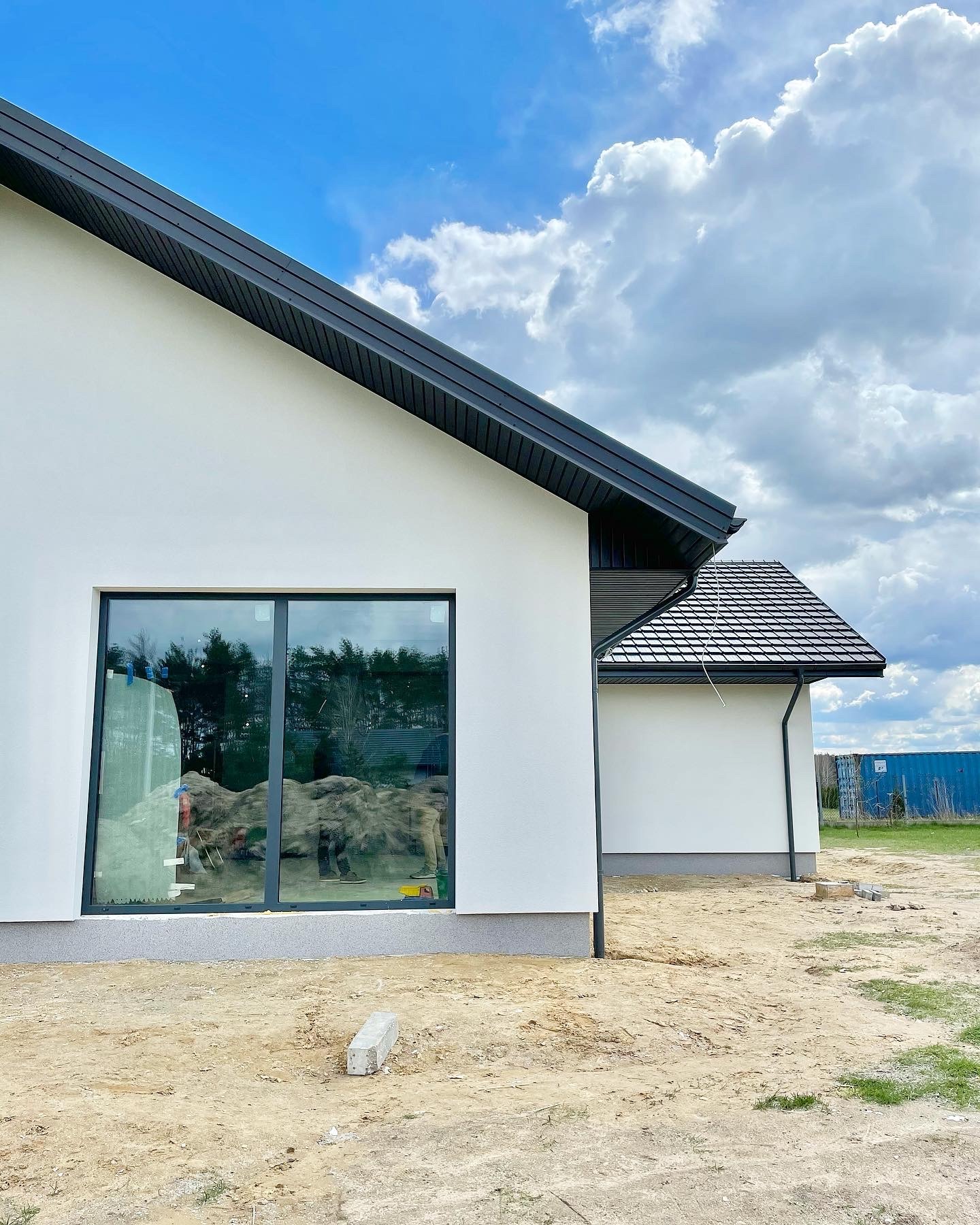 Fragment nowoczesnego, białego domu z czarnym dachem i oknami, widoczne przygotowane podłoże i odblask w oknie.