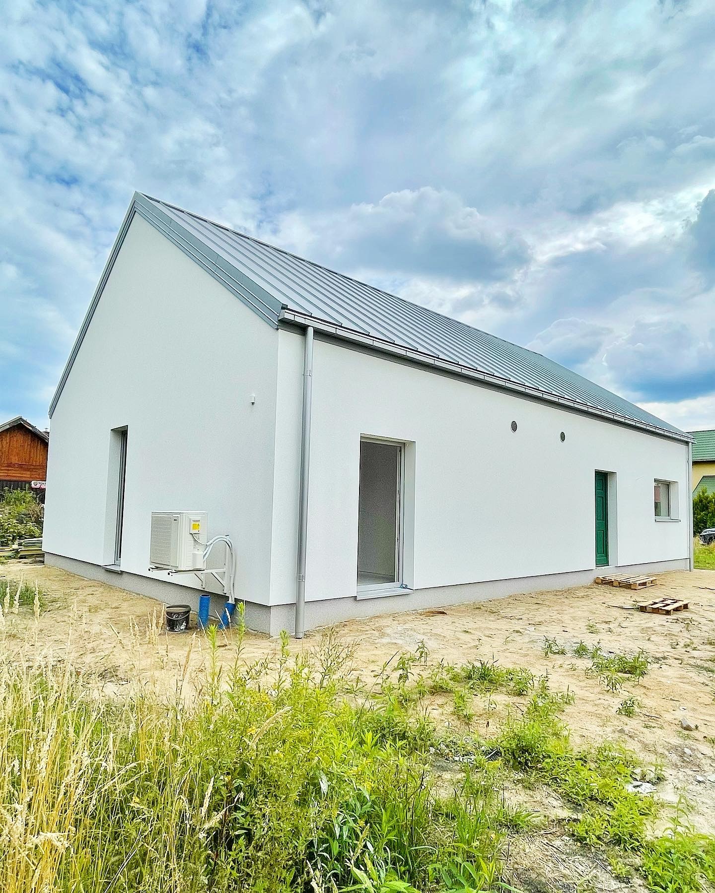 Nowoczesny, parterowy dom z białymi ścianami i szarym dachem, z klimatyzacją zewnętrzną, widok z zewnątrz na trawiastą działkę z chmurami na niebie.