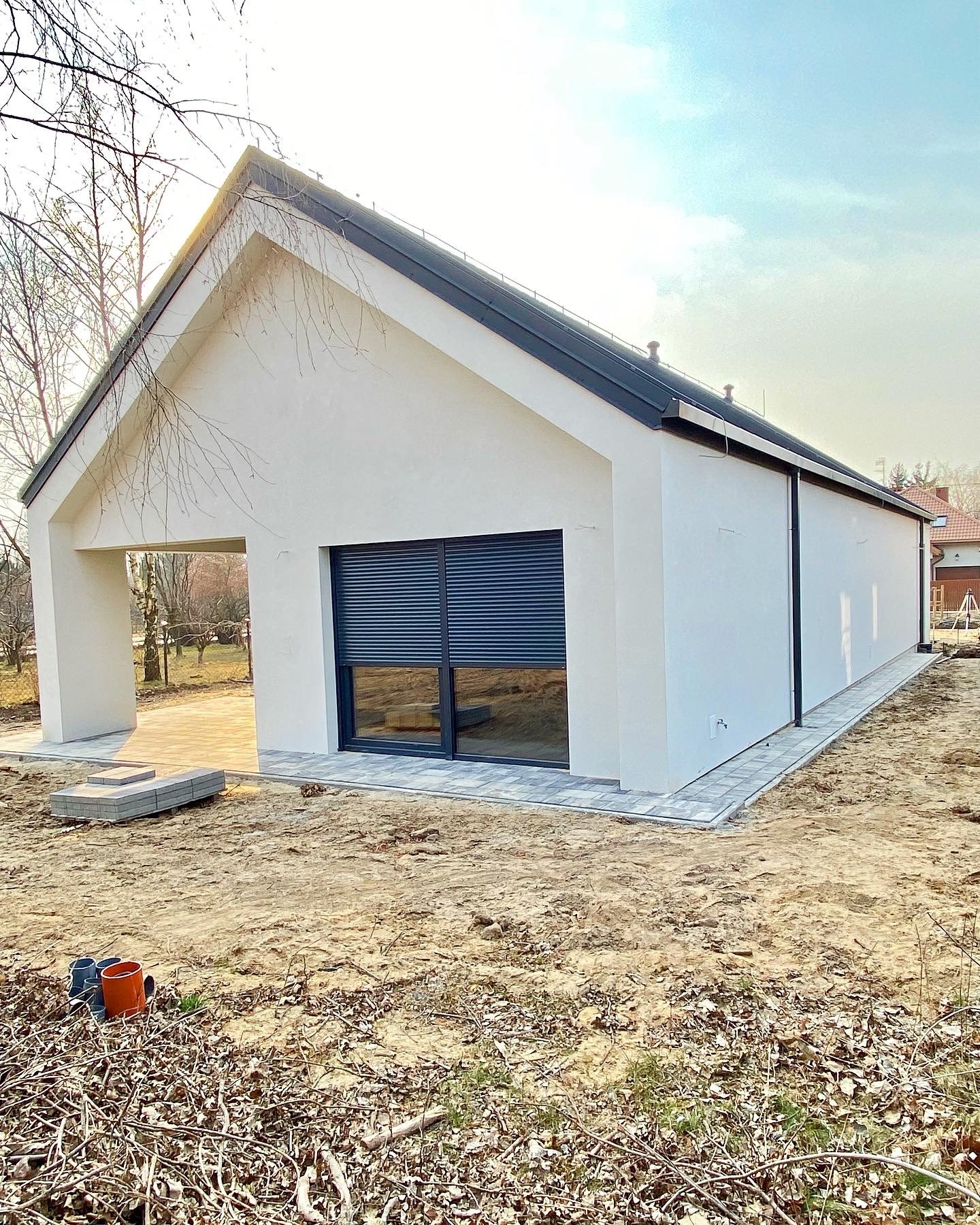 Nowoczesny, parterowy dom z białym tynkiem, grafitową stolarką okienną i dachem, widok z zewnątrz na częściowo wykończony taras i posesję w stanie surowym.