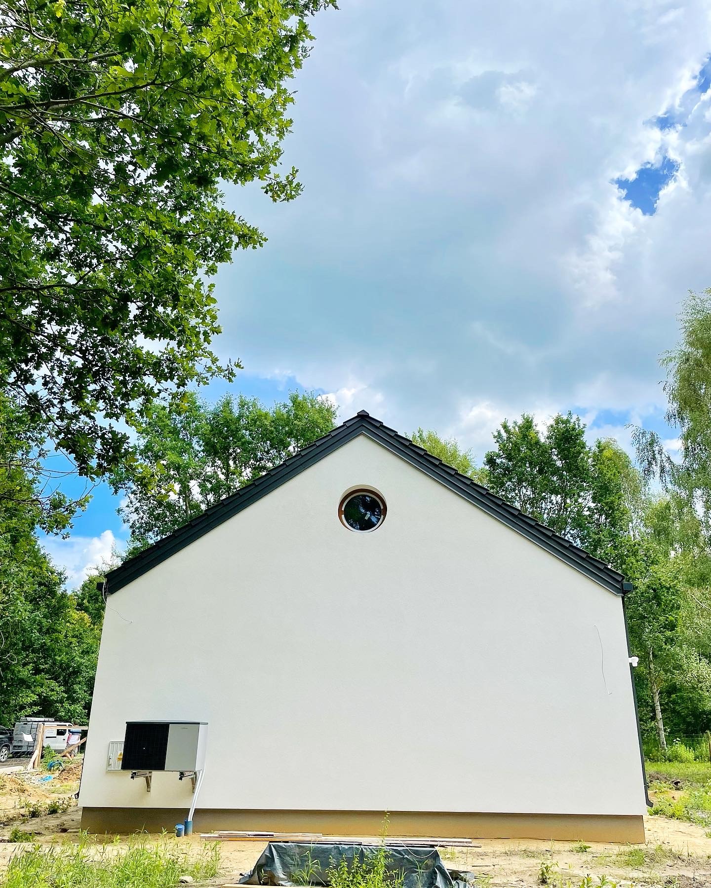 Nowoczesny dom jednorodzinny z białymi ścianami, okrągłym oknem na szczycie i ciemnym dachem, otoczony zielenią drzew, z widoczną pompą ciepła na bocznej ścianie.
