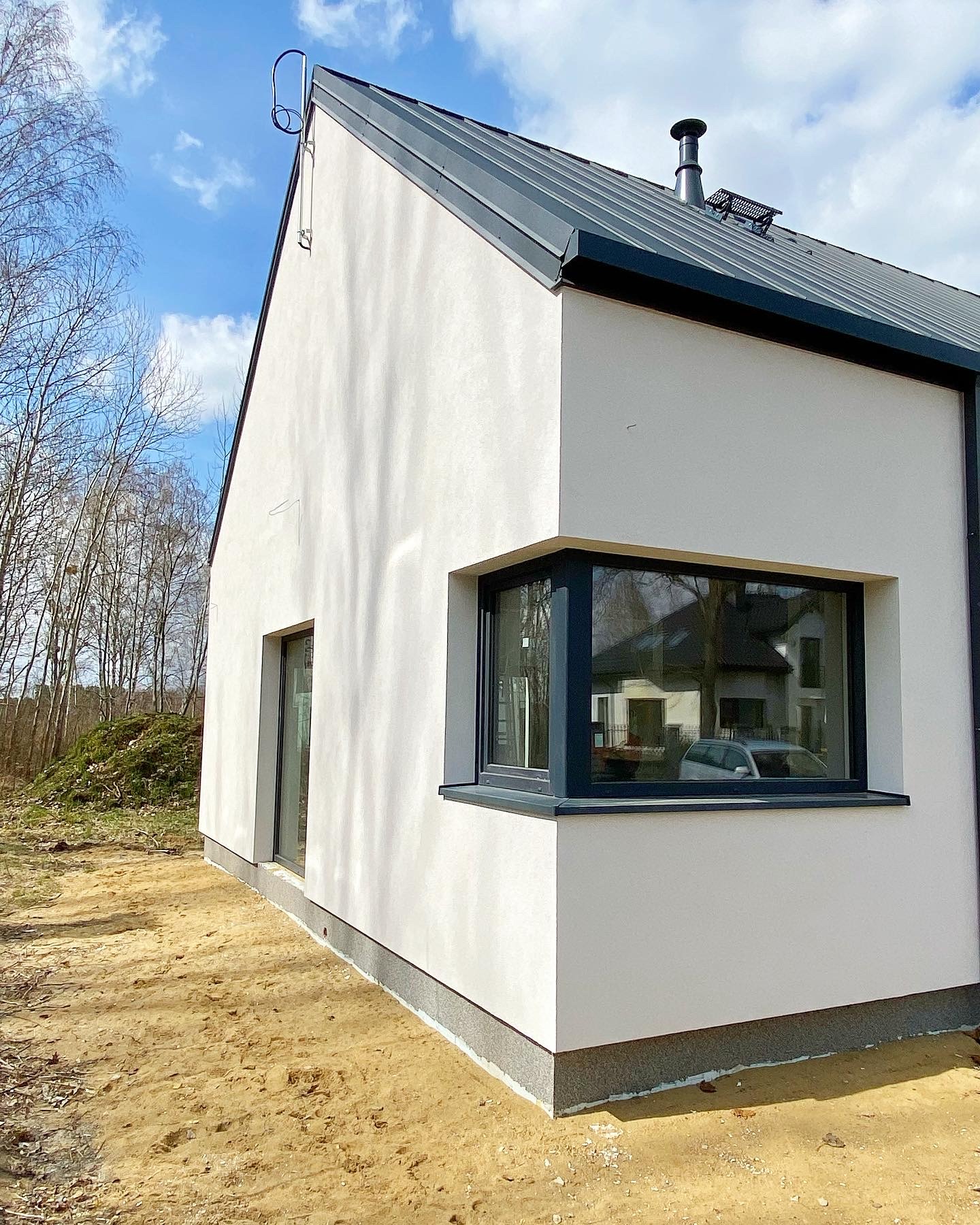 Nowoczesny, biały dom jednorodzinny z ciemną, metalową dachówką i dużym narożnym oknem, widok z zewnątrz na niezagospodarowany jeszcze teren.