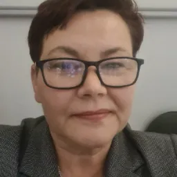 Portret kobiety w okularach i szarym żakiecie, z kr&oacute;tkimi brązowymi włosami, patrzącej w obiektyw, z odbiciami w szkłach okular&oacute;w.