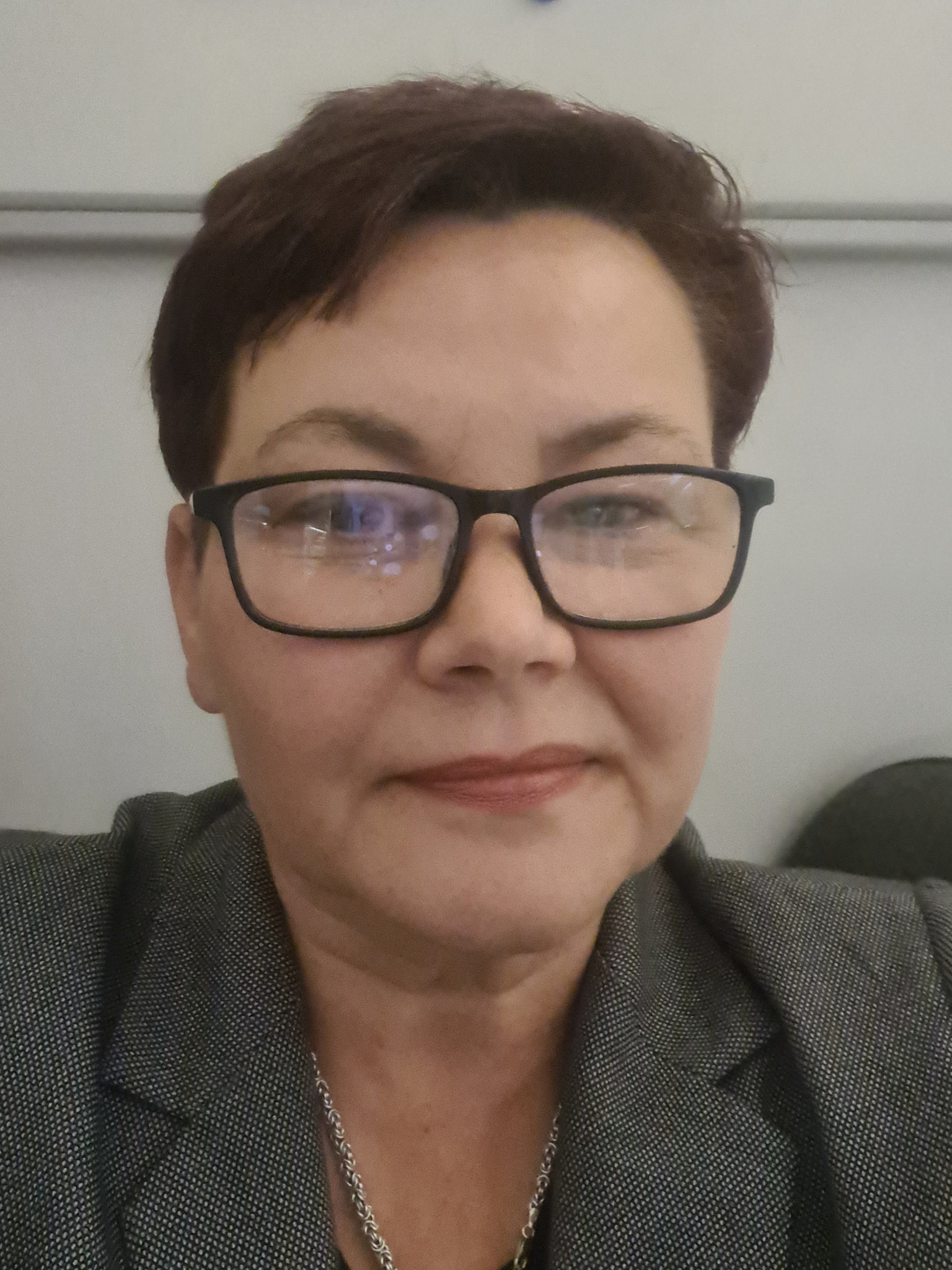 Portret kobiety w okularach i szarym żakiecie, z krótkimi brązowymi włosami, patrzącej w obiektyw, z odbiciami w szkłach okularów.