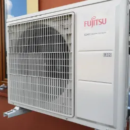 Zewnętrzna jednostka klimatyzacji Fujitsu DC Inverter R32, zamontowana na brązowej ścianie budynku, widoczny fragment okna z odbiciem nieba.