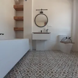 Nowoczesna łazienka po remoncie z wzorzystą, kremowo-brązową podłogą, białą wanną, umywalką z szafką i okrągłym lustrem podświetlanym lampą, oraz toaletą z drewnianą deską i dekoracyjną lampą...