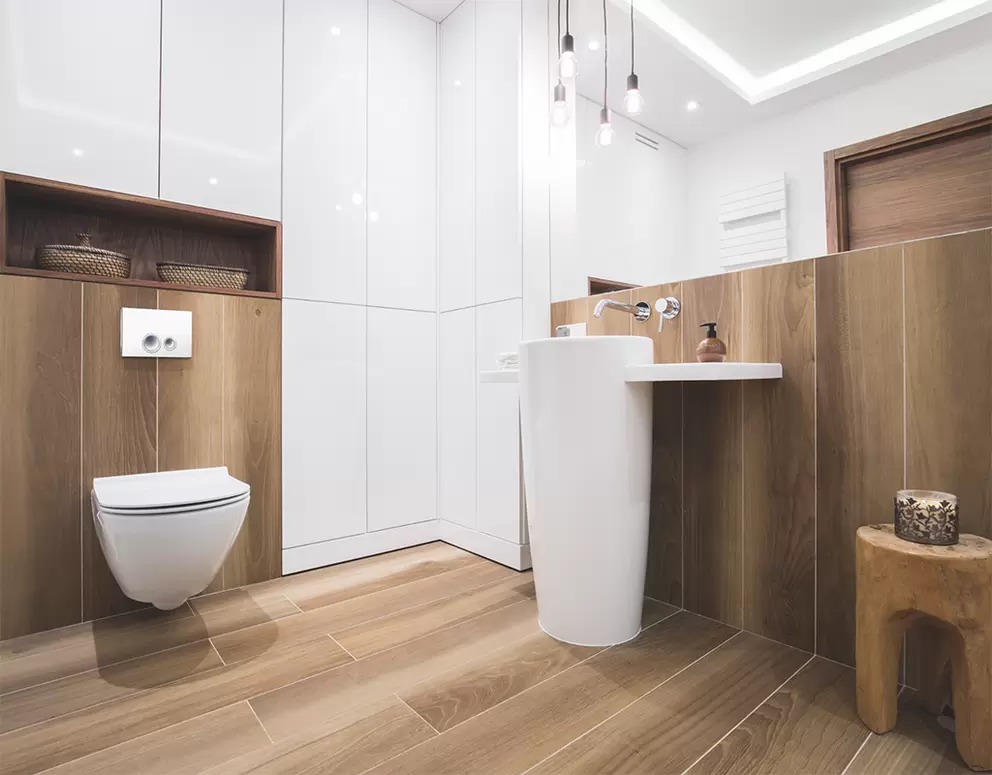 Nowoczesna łazienka z drewnopodobnymi płytkami na podłodze i ścianach, wisząca toaleta, umywalka na postumencie i drewniany stołek.