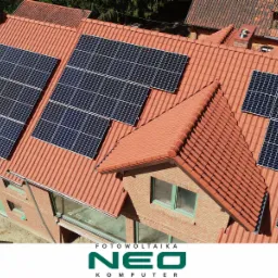 Widok z góry na dom z czerwoną dachówką, na której zamontowane są panele słoneczne, z widocznym logo firmy na dole kadru.
