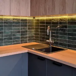 Nowoczesna kuchnia z ciemnozielonymi, podłużnymi płytkami na ścianie, podświetleniem LED pod szafkami, drewnianym blatem i czarnym zlewozmywakiem z chromowaną baterią.