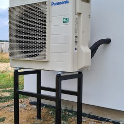 Ergo-klima Tomasz Menzel - Zewnętrzna jednostka klimatyzacji Panasonic R32 zamontowana na czarnym metalowym stelażu przy białej ścianie budynku, z widocznymi przewodami w izolacji wchodzącymi do otworu w ścianie.