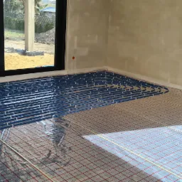 Niebieskie rury ogrzewania podłogowego rozłożone na folii z siatką w surowym wnętrzu domu w trakcie budowy, widok z góry, naturalne światło słoneczne.