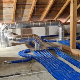 Ergo-klima Tomasz Menzel - Instalacja przewodów wentylacyjnych z niebieskiego tworzywa sztucznego i metalowych rur w surowym wnętrzu poddasza z drewnianą konstrukcją dachu i ścianami z betonu komórkowego.