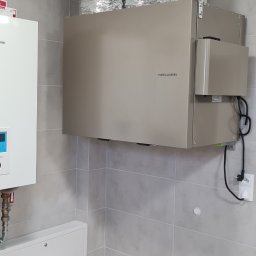 Ergo-klima Tomasz Menzel - Zamontowany rekuperator Thessla Green z trzema srebrnymi, elastycznymi przewodami wentylacyjnymi w pomieszczeniu z szarymi płytkami, widoczny biały piec po lewej stronie.